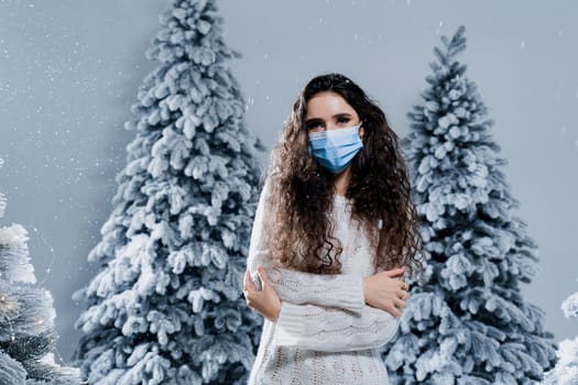 New year celebration at covid-19 coronavirus quarantine period. Happy girl in medical mask with falling snow stays at home. Social distance. Winter holidays in snowy day