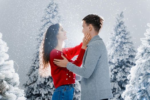 Happy young couple hugs and kiss near christmas trees at the eve of new year celebration in winter day. Smiley man and woman love each other. Falling snow and kisses.