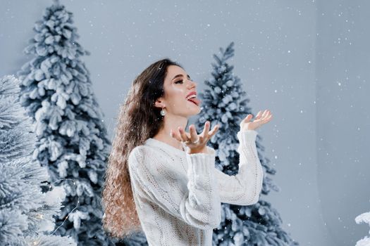 Happy girl smiles and touches falling snow at eve new year. Young woman near big snowy spruces in winter day before christmas celebration