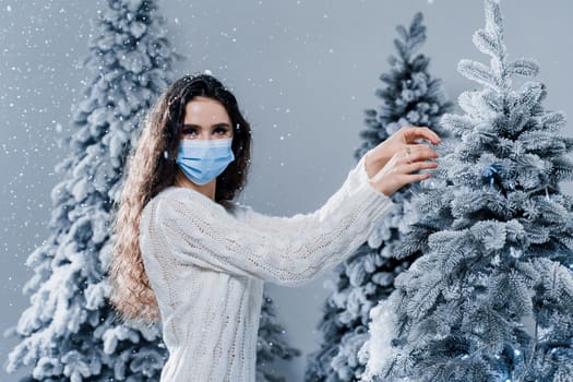 New year celebration at covid-19 coronavirus quarantine period. Happy girl in medical mask with falling snow stays at home. Social distance. Winter holidays in snowy day