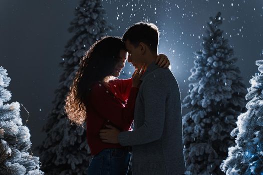 Falling snow and kisses with falling snow on dark blue background. Happy young couple hug and kiss near christmas trees at the eve of new year celebration in winter day