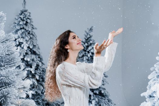 Happy girl smiles and touches falling snow at eve new year. Young woman near big snowy spruces in winter day before christmas celebration