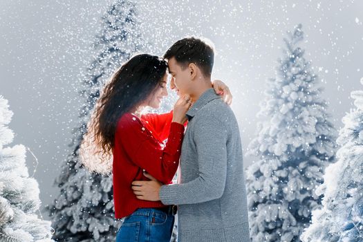 Happy young couple hugs and kiss near christmas trees at the eve of new year celebration in winter day. Smiley man and woman love each other. Falling snow and kisses.