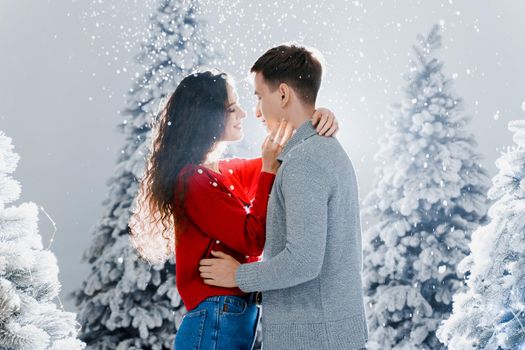 Happy young couple hugs and kiss near christmas trees at the eve of new year celebration in winter day. Smiley man and woman love each other. Falling snow and kisses.