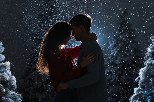 Falling snow and kisses with falling snow on dark blue background. Happy young couple hug and kiss near christmas trees at the eve of new year celebration in winter day