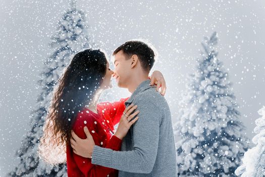 Happy young couple hugs and kiss near christmas trees at the eve of new year celebration in winter day. Smiley man and woman love each other. Falling snow and kisses.