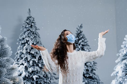 New year celebration at covid-19 coronavirus quarantine period. Happy girl in medical mask with falling snow stays at home. Social distance. Winter holidays in snowy day