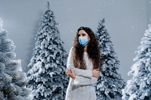 New year celebration at covid-19 coronavirus quarantine period. Happy girl in medical mask with falling snow stays at home. Social distance. Winter holidays in snowy day