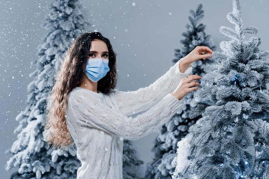 New year celebration at covid-19 coronavirus quarantine period. Happy girl in medical mask with falling snow stays at home. Social distance. Winter holidays in snowy day