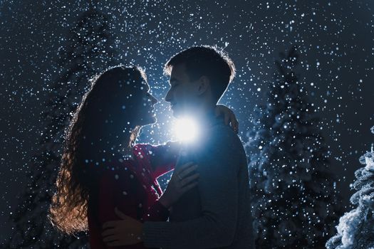 Falling snow and kisses with falling snow on dark blue background. Happy young couple hug and kiss near christmas trees at the eve of new year celebration in winter day
