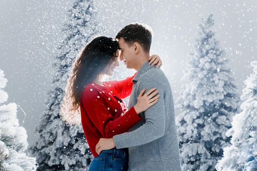 Happy young couple hugs and kiss near christmas trees at the eve of new year celebration in winter day. Smiley man and woman love each other. Falling snow and kisses.