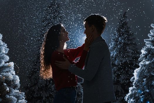 Falling snow and kisses with falling snow on dark blue background. Happy young couple hug and kiss near christmas trees at the eve of new year celebration in winter day