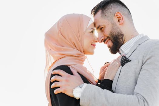 Muslim love story close-up with sun light. Mixed couple smiles and hugs at sunset. Woman weared in hijab looks to her man. Advert for on-line dating agency