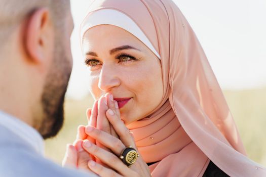 Muslim love story close-up. Mixed couple smiles and hugs on the green hills . Woman weared in hijab looks to her man. Advert for on-line dating agency