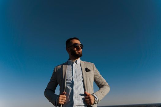 Handsome bearded arabian man in sunglasses on the blue sky background. Confident muslim business man. Arab man weared in grey coat