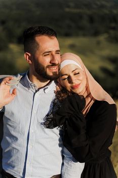 Muslim love story close-up. Mixed couple smiles and hugs on the green hills . Woman weared in hijab looks to her man. Advert for on-line dating agency