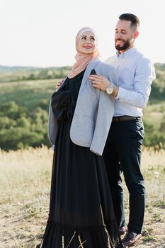 Muslim love story close-up. Mixed couple smiles and hugs on the green hills . Woman weared in hijab looks to her man. Advert for on-line dating agency