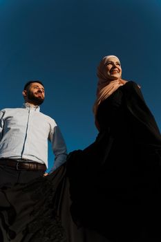 Muslim love story on the blue sky background. cheerful mixed couple smiles and hugs . Woman weared in hijab looks to her man. Advert for on-line dating agency