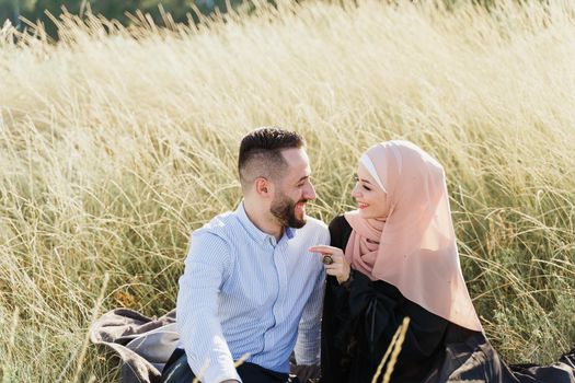 Muslim love story. Mixed couple seats on the grass, smiles and hugs.. Woman weared in hijab looks to her man. Advert for on-line dating agency
