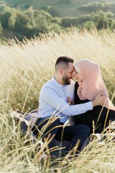 Muslim love story. Mixed couple seats on the grass, smiles and hugs.. Woman weared in hijab looks to her man. Advert for on-line dating agency