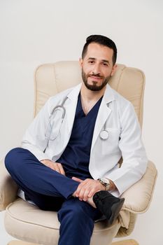 Arabian doctor surgeon in medical robe with phonendoscope seats in armchair in studio on white blanked background. Confident arab on white background