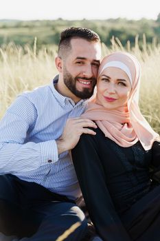 Muslim love story close-up. Mixed couple smiles and hugs on the green hills . Woman weared in hijab looks to her man. Advert for on-line dating agency