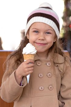 Portrait of happy and smiling caucasian little girl of 5-6 years holding ice cream cone outside in light brown coat and hat