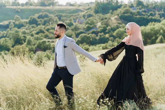 Muslim love story of mixed couple. Man and woman smiles and walks on the green hills . Woman weared in hijab looks to her man. Advert for on-line dating agency