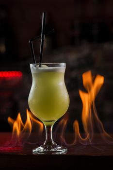 Bartender sprinkles on illuminated glass with bright green cold cocktail on bar counter and makes fire flame over it.