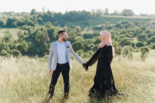 Muslim love story of mixed couple. Man and woman smiles and walks on the green hills . Woman weared in hijab looks to her man. Advert for on-line dating agency