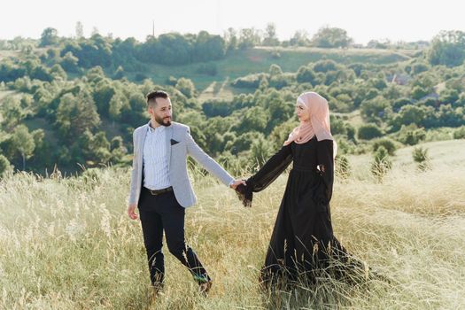 Muslim love story of mixed couple. Man and woman smiles and walks on the green hills . Woman weared in hijab looks to her man. Advert for on-line dating agency