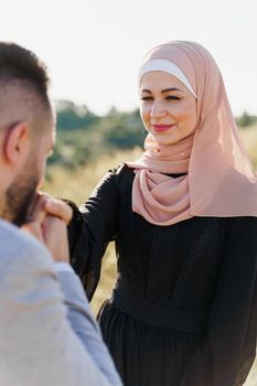 Muslim love story close-up. Mixed couple smiles and hugs on the green hills . Woman weared in hijab looks to her man. Advert for on-line dating agency