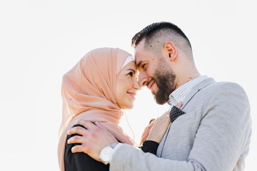 Muslim love story close-up with sun light. Mixed couple smiles and hugs at sunset. Woman weared in hijab looks to her man. Advert for on-line dating agency