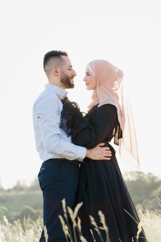 Muslim love story with sun light. Mixed couple smiles and hugs at sunset. Woman weared in hijab looks to her man. Advert for on-line dating agency