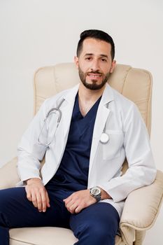Arabian doctor surgeon in medical robe with phonendoscope seats in armchair in studio on white blanked background. Confident arab on white background