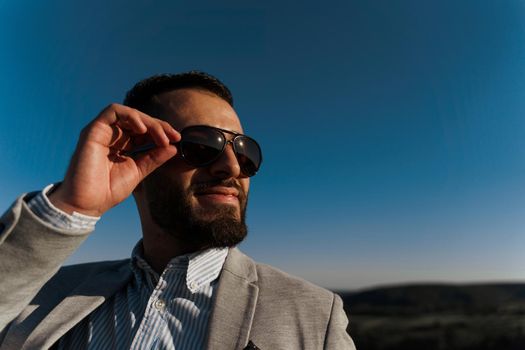 Handsome bearded arabian man in sunglasses on the blue sky background. Confident muslim business man. Arab man weared in grey coat