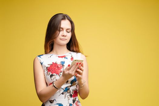 Front view of pretty female in bright dress keeping phone and chattering. Attractive girl messaging and using gadget on yellow isolated background in studio. Concept of communication.