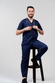 Handsome arab doctor surgeon smiles on white background. Bearded student with stethoscope in medical robe seats on chair on blanked background.