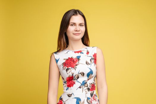 Studio portrait of fashionable brunette lady in white dress with flowers and beige heels posing with bent leg. Smiling at camera. holding skirt. Isolate on yellow background.