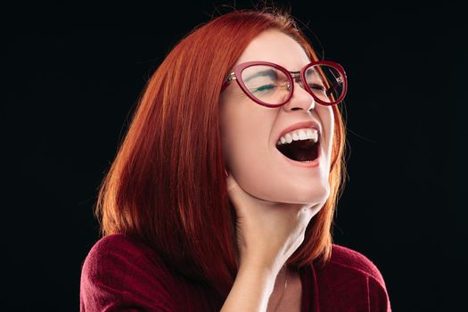 Ill young woman with closed eyes and opened mouth shouting because of her sore throat. Girl with ginger hair having pain in neck or throat. Beautiful model wearing spectacles.