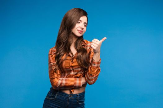 Front view of beautiful woman wearing checkered shirt looking aside and thinking on blue isolated background. Young long haired girl keeping fingers near chin and dreaming. Concept of workday.