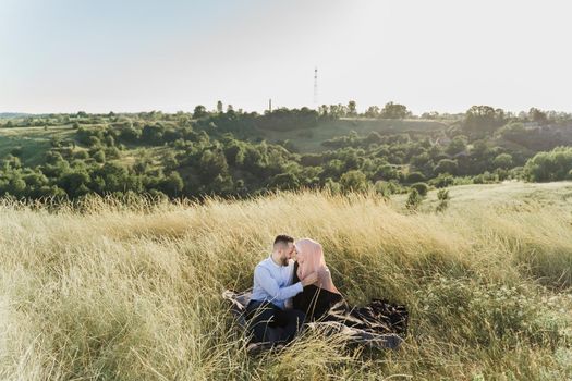 Muslim love story. Mixed couple seats on the grass, smiles and hugs.. Woman weared in hijab looks to her man. Advert for on-line dating agency