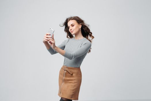 Full length portrait of happy stylish woman with charming smile in fashion sweater and brown skirt. She is standing and taking selfie with smartphone. Isolated on gray background
