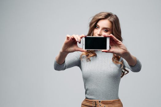 Waist up portrait of attractive girl holding modern smartphone in hands and taking selfie. Cheerful female with long brown hair and lovely eyes in fashionable sweater. Isolated on gray background