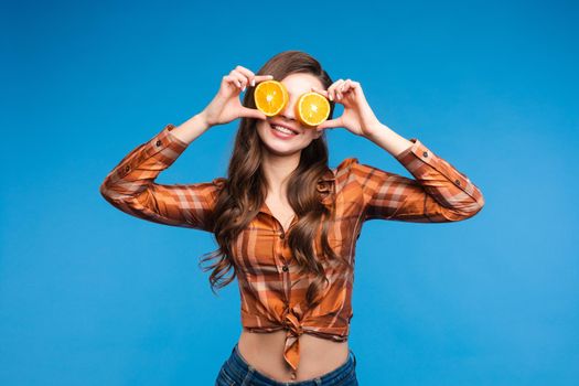 Young girl in casual clothes hiding her face behind fruits. Brunette lady in checkered shirt and jeans holding halves of oranges as sunglasses. Beatiful cute woman with opened belly happily smiling.