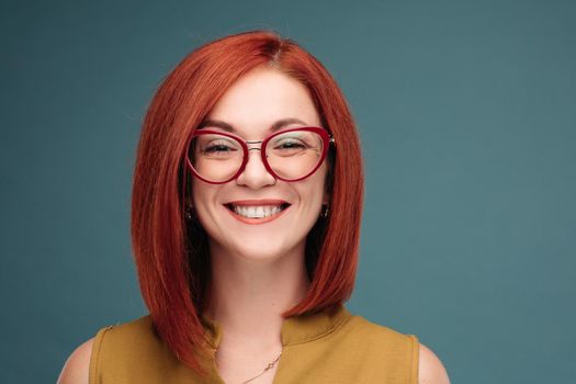 Studio portrait of a happy girl with brown hair. A red-haired woman in stylish glasses smiles looking at the camera. Close-up. Isolated on a blue background