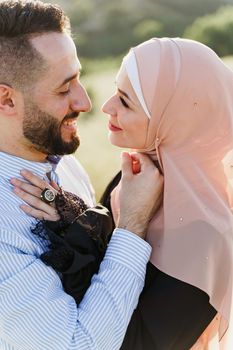 Muslim love story close-up. Mixed couple smiles and hugs on the green hills . Woman weared in hijab looks to her man. Advert for on-line dating agency