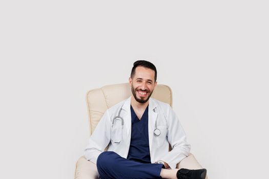 Handsome arab doctor surgeon smiles on white background. Bearded student with stethoscope in medical robe seats in armchair on blanked background.