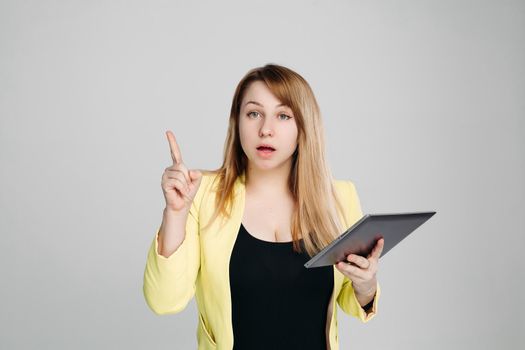 Portrait of creative blonde haired woman, wearing in yellow jacket and black blouse, holding tablet computer and pointing finger up against white studio background. Concept of business ideas.