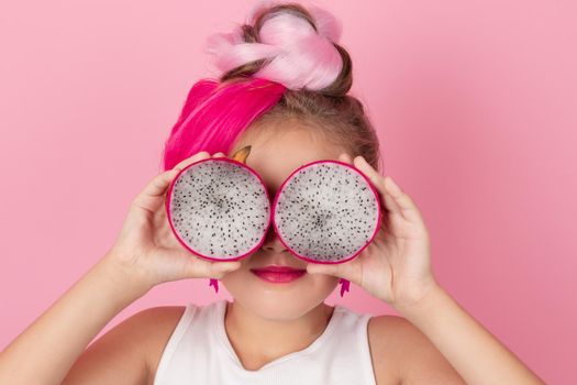 Close-up portrait of pretty girl with pink hairstyle with dragon fruit on pink background. Studio shot of charming tween girl with pink make up enjoying juicy red pitaya. exotic Pitahaya fruit.